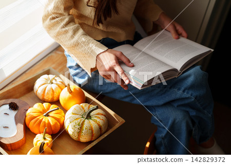 Teen girl reading book by window with pumpkins in autumn light Teen girl reading book by window with pumpkins in autumn light 142829372