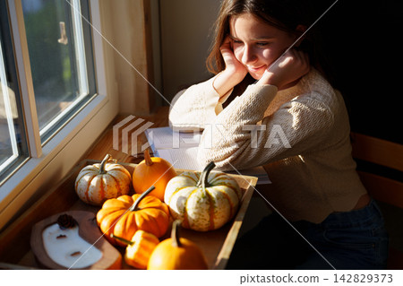 Teen girl relaxing by window with decorative pumpkins at home Teen girl relaxing by window with decorative pumpkins at home 142829373