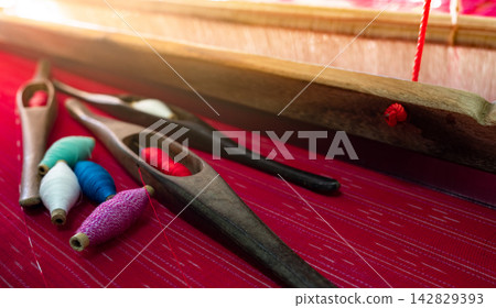 Traditional wooden weaving shuttle and colorful thread on red handmade fabric, textile loom tools used by artisan for cloth production, cultural craftsmanship and heritage weaving technique. Traditional wooden weaving shuttle and colorful thread on red handmade fabric, textile loom tools used by artisan for cloth production, cultural craftsmanship and heritage weaving technique. 142829393