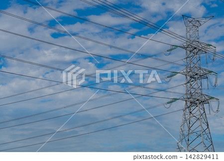 High voltage electricity transmission tower and power lines against blue cloudy sky, electrical grid infrastructure delivering energy for urban communities, industry and modern power distribution. High voltage electricity transmission tower and power lines against blue cloudy sky, electrical grid infrastructure delivering energy for urban communities, industry and modern power distribution. 142829411