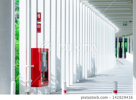 Red fire extinguisher cabinet along an empty modern building corridor with repeating white columns and sunlight shadows, providing emergency fire safety equipment for public building protection. Red fire extinguisher cabinet along an empty modern building corridor with repeating white columns and sunlight shadows, providing emergency fire safety equipment for public building protection. 142829416