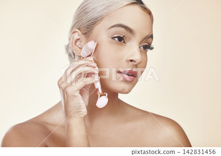 Facial roller, skincare and woman in studio with natural, health and cosmetic routine. Beauty, wellness and female person with rose quartz dermatology tool for face treatment by white background. Facial roller, skincare and woman in studio with natural, health and cosmetic routine. Beauty, wellness and female person with rose quartz dermatology tool for face treatment by white background. 142831456