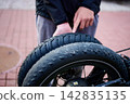 A person is pointing at a bicycle tire while holding it in a city setting. The scene captures a moment of maintenance or inspection 142835135