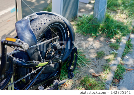 A bicycle is parked on a sidewalk with a flat rear tire. Grass grows nearby under clear skies. The setting is typical for an urban area A bicycle is parked on a sidewalk with a flat rear tire. Grass grows nearby under clear skies. The setting is typical for an urban area 142835141
