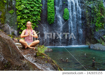 Young woman meditating in yoga pose under Banyumala Twin Waterfalls. Water flowing under cascading high waterfall. Adventure lifestyle and retreat at holidays travel in Bali tropical rainforest. Young woman meditating in yoga pose under Banyumala Twin Waterfalls. Water flowing under cascading high waterfall. Adventure lifestyle and retreat at holidays travel in Bali tropical rainforest. 142835155