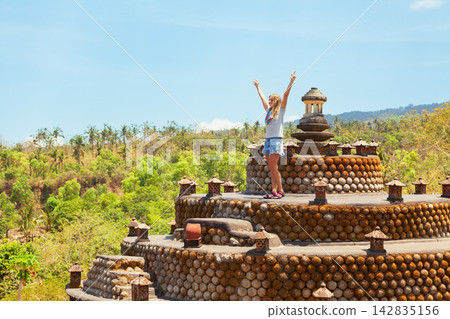 Young joyful woman stands with raised hands on buddhist stupa terrace at Aung mount slopes in Tulamben, Bali, Indonesia. Eco landmark inspired by Borobudur, surrounded by tropical forest and blue sky. Young joyful woman stands with raised hands on buddhist stupa terrace at Aung mount slopes in Tulamben, Bali, Indonesia. Eco landmark inspired by Borobudur, surrounded by tropical forest and blue sky. 142835156