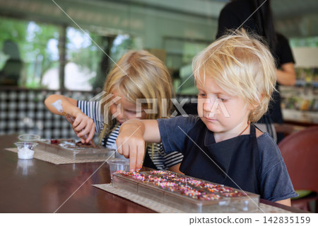 Two young children concentrate on decorating handmade chocolate with colorful toppings during a fun workshop at a Bali. A sweet and educational travel activity for families. Two young children concentrate on decorating handmade chocolate with colorful toppings during a fun workshop at a Bali. A sweet and educational travel activity for families. 142835159
