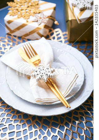 Christmas festive table setting with white plates and gold cutlery, gifts in white and gold paper, decorations and balls. Christmas festive table setting with white plates and gold cutlery, gifts in white and gold paper, decorations and balls. 142835403