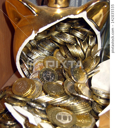Close-up of a broken golden piggy bank with a crown spilling numerous metal coins, representing wealth, savings, and financial investment. Close-up of a broken golden piggy bank with a crown spilling numerous metal coins, representing wealth, savings, and financial investment. 142835535