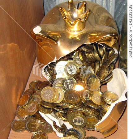 Broken golden piggy bank with a crown overflowing with metallic coins on a wooden shelf, symbolizing savings, wealth, and financial concepts. Broken golden piggy bank with a crown overflowing with metallic coins on a wooden shelf, symbolizing savings, wealth, and financial concepts. 142835538