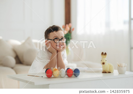 Young boy with glasses smiling while resting his chin on hands, surrounded by colorful decorative eggs and a small toy deer on a white table in a bright room 142835975