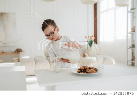 Young boy with glasses pouring milk from a glass pitcher into a clear glass beside a plate of chocolate chip cookies in a bright, modern kitchen setting Young boy with glasses pouring milk from a glass pitcher into a clear glass beside a plate of chocolate chip cookies in a bright, modern kitchen setting 142835976