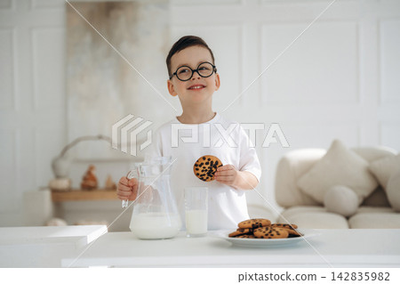 Young boy with glasses holds cookie and smiles while standing next to a glass of milk and a plate of cookies in a bright, modern kitchen setting 142835982