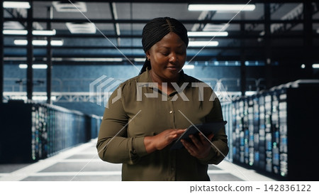 Data center engineer monitors data center metrics on tablet to enhance rigs operational effectiveness. Worker walks through server farm rows, using analytics on device to predict performance, camera B Data center engineer monitors data center metrics on tablet to enhance rigs operational effectiveness. Worker walks through server farm rows, using analytics on device to predict performance, camera B 142836122