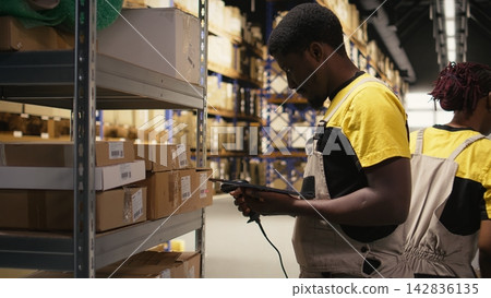 African American male employee scanning packages in a large warehouse, registering airway bills shipment details within industrial depot records. Using scanner for order fulfillment. Camera A. African American male employee scanning packages in a large warehouse, registering airway bills shipment details within industrial depot records. Using scanner for order fulfillment. Camera A. 142836135