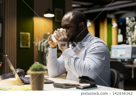 African american male analyst drinking coffee cup to keep working at night, staying awake to finish administrative tasks in the office. Sipping on a caffeine drink after hours. African american male analyst drinking coffee cup to keep working at night, staying awake to finish administrative tasks in the office. Sipping on a caffeine drink after hours. 142836162