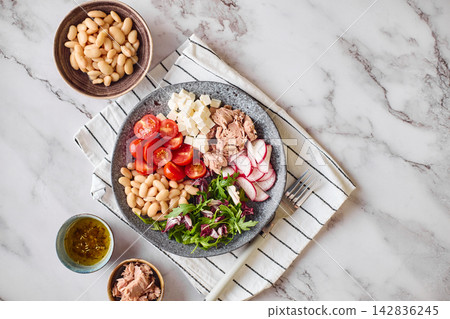 Healthy tuna salad bowl with white beans, feta cheese, tomatoes, radish, and greens on marble table Healthy tuna salad bowl with white beans, feta cheese, tomatoes, radish, and greens on marble table 142836245