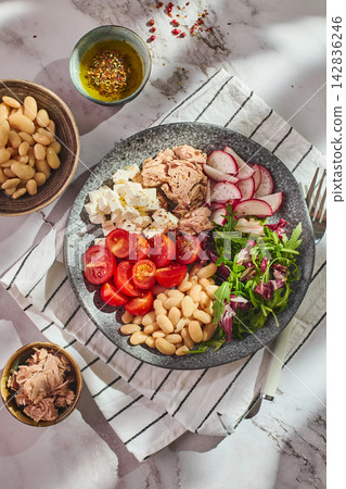 Healthy tuna salad bowl with white beans, feta cheese, tomatoes, radish, and greens on marble table Healthy tuna salad bowl with white beans, feta cheese, tomatoes, radish, and greens on marble table 142836246