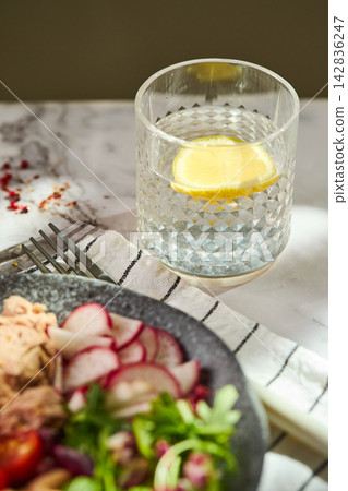 Close up of crystal glass with water and lemon slice next to healthy salad bowl on marble table Close up of crystal glass with water and lemon slice next to healthy salad bowl on marble table 142836247