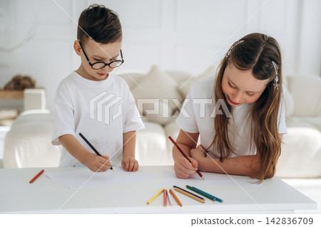 Young children engaged in creative drawing activity with colored pencils on a white table in a bright, modern living room with soft furnishings and natural light Young children engaged in creative drawing activity with colored pencils on a white table in a bright, modern living room with soft furnishings and natural light 142836709