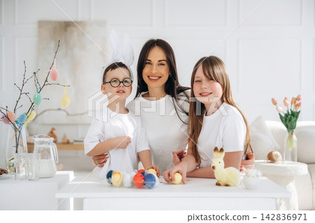 Family of three, including a woman and two children, smiling together at a table decorated with colorful eggs and Easter-themed items in a bright indoor setting Family of three, including a woman and two children, smiling together at a table decorated with colorful eggs and Easter-themed items in a bright indoor setting 142836971