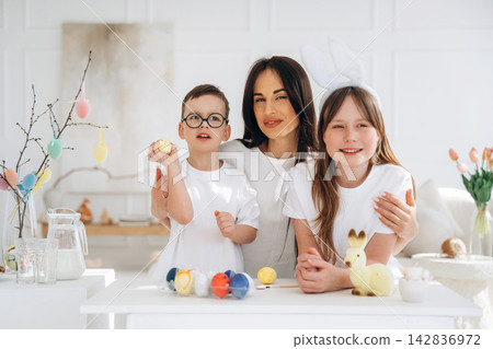 Family celebrating Easter with colorful eggs and decorations, mother and two children smiling together at a bright, modern table in a well-lit room Family celebrating Easter with colorful eggs and decorations, mother and two children smiling together at a bright, modern table in a well-lit room 142836972