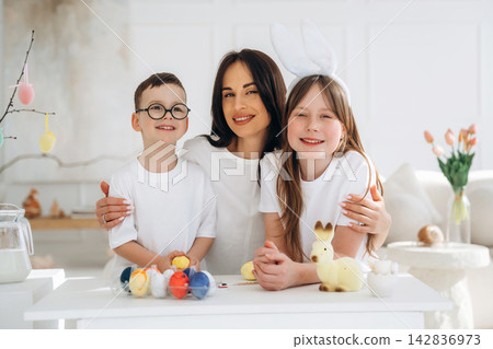 Woman with two children wearing white shirts and bunny ears, celebrating Easter with colorful eggs and decorations in a bright, modern living room Woman with two children wearing white shirts and bunny ears, celebrating Easter with colorful eggs and decorations in a bright, modern living room 142836973