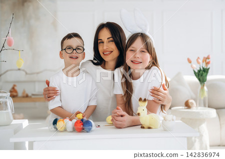 Caucasian woman with children wearing white outfits and bunny ears, celebrating Easter with colorful eggs and decorations in a bright, modern living room Caucasian woman with children wearing white outfits and bunny ears, celebrating Easter with colorful eggs and decorations in a bright, modern living room 142836974