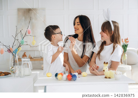 Family celebrating Easter with colorful eggs and decorations, mother and children smiling while preparing festive treats in a bright, modern kitchen Family celebrating Easter with colorful eggs and decorations, mother and children smiling while preparing festive treats in a bright, modern kitchen 142836975