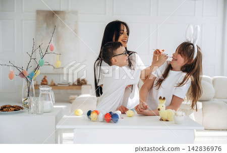 Family celebrating Easter with children painting eggs and decorating a table with colorful ornaments and treats in a bright, modern living room setting Family celebrating Easter with children painting eggs and decorating a table with colorful ornaments and treats in a bright, modern living room setting 142836976
