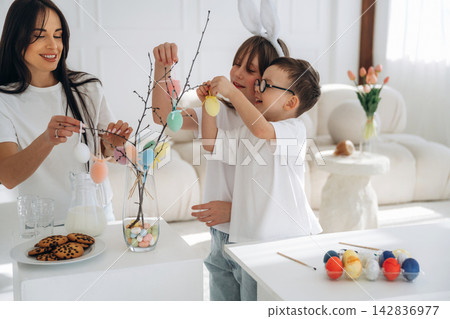 Woman and two children decorating a branch with colorful Easter eggs in a bright, modern living room with cookies and milk on a white table Woman and two children decorating a branch with colorful Easter eggs in a bright, modern living room with cookies and milk on a white table 142836977