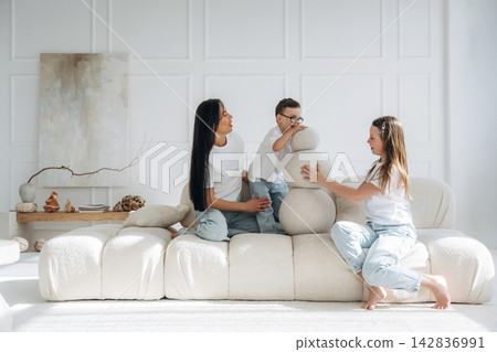 Two women and a boy play with a soft sculpture on a modern white sofa in a bright, minimalist living room with natural light and decorative elements Two women and a boy play with a soft sculpture on a modern white sofa in a bright, minimalist living room with natural light and decorative elements 142836991