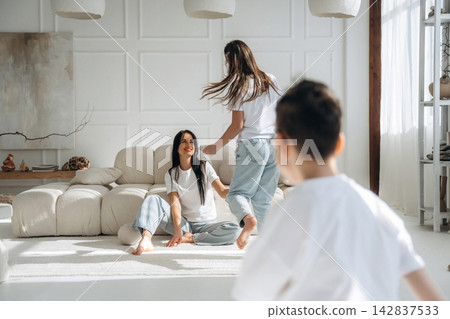 Two women and a boy play together in a bright, modern living room with a white sofa, light wood accents, and large windows allowing natural light to fill the space Two women and a boy play together in a bright, modern living room with a white sofa, light wood accents, and large windows allowing natural light to fill the space 142837533