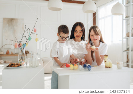 Family of three, including a woman and two children, decorating colorful Easter eggs at a white table in a bright, modern living room with festive decorations Family of three, including a woman and two children, decorating colorful Easter eggs at a white table in a bright, modern living room with festive decorations 142837534