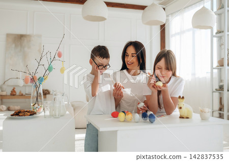 Family decorating Easter eggs together in a bright, modern living room with a decorative branch and colorful eggs on a white table Family decorating Easter eggs together in a bright, modern living room with a decorative branch and colorful eggs on a white table 142837535