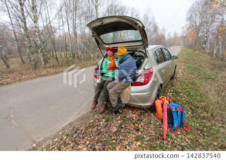 An elderly couple enjoys snacks while sitting by a parked car on the side of the road surrounded by trees in autumn. An elderly couple enjoys snacks while sitting by a parked car on the side of the road surrounded by trees in autumn. 142837540