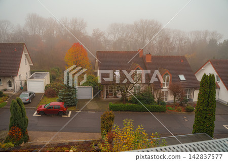 Foggy autumn residential neighborhood with suburban houses. Quiet residential neighborhood, detached houses, autumn trees, parked cars covered by soft morning fog, magic suburban environment in fall Foggy autumn residential neighborhood with suburban houses. Quiet residential neighborhood, detached houses, autumn trees, parked cars covered by soft morning fog, magic suburban environment in fall 142837577