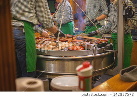 Sausage vendors grill sausage varieties at Christmas city market, serving food to visitors. Street food service, market catering, holiday tourism, retail and seasonal dining, busy market or festival Sausage vendors grill sausage varieties at Christmas city market, serving food to visitors. Street food service, market catering, holiday tourism, retail and seasonal dining, busy market or festival 142837578