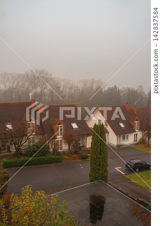 Residential houses along street under dense fog with trees and tiled roofs. Houses with red tiled roofs, autumn vegetation, quiet street fading into dense morning fog, calm residential fall scene Residential houses along street under dense fog with trees and tiled roofs. Houses with red tiled roofs, autumn vegetation, quiet street fading into dense morning fog, calm residential fall scene 142837584
