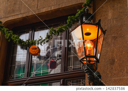 Vintage street lamp glowing beside historic building. Ornate vintage street lamp glowing, warm orange light beside historic stone building, decorative window, atmospheric urban architecture background Vintage street lamp glowing beside historic building. Ornate vintage street lamp glowing, warm orange light beside historic stone building, decorative window, atmospheric urban architecture background 142837586