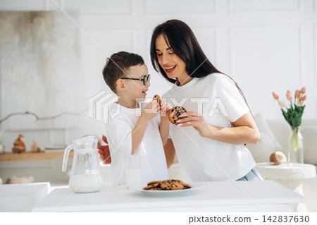 Woman and boy in white shirts share chocolate chip cookies in a bright kitchen, with a pitcher of milk and a plate of cookies on the table 142837630