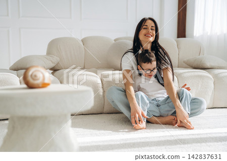 Woman with long dark hair sitting on a light carpeted floor, playing with a young boy in glasses, both smiling in a bright, modern living room with a beige sofa 142837631