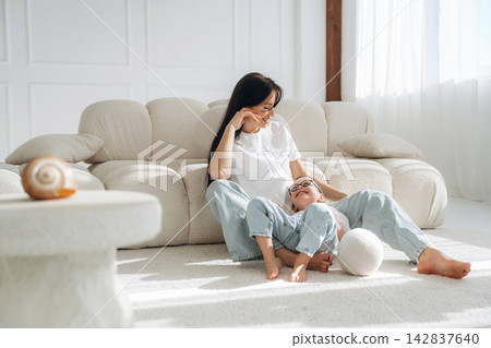 Woman and child sitting on a light-colored sofa in a bright living room, enjoying a playful moment together on a soft carpet with natural light streaming in Woman and child sitting on a light-colored sofa in a bright living room, enjoying a playful moment together on a soft carpet with natural light streaming in 142837640
