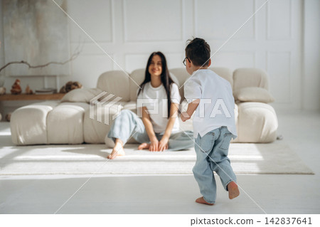 Young boy in casual clothing runs towards smiling woman seated on a light-colored sofa in a bright, modern living room with minimalistic decor Young boy in casual clothing runs towards smiling woman seated on a light-colored sofa in a bright, modern living room with minimalistic decor 142837641