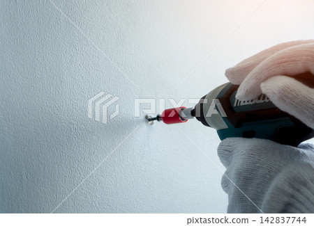 Gloved hands using cordless drill to drive screw into white interior wall, home renovation work for mounting hardware during DIY repair, installation and apartment improvement project. Gloved hands using cordless drill to drive screw into white interior wall, home renovation work for mounting hardware during DIY repair, installation and apartment improvement project. 142837744