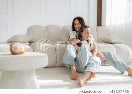 Two women sitting on a light-colored sofa in a bright living room, one woman embracing the other, with a decorative shell on a table nearby Two women sitting on a light-colored sofa in a bright living room, one woman embracing the other, with a decorative shell on a table nearby 142837786