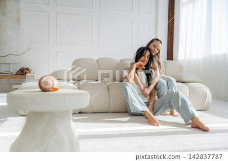 Two women sitting on a light-colored sofa in a modern living room, with natural light streaming through large windows and a decorative table in the foreground Two women sitting on a light-colored sofa in a modern living room, with natural light streaming through large windows and a decorative table in the foreground 142837787