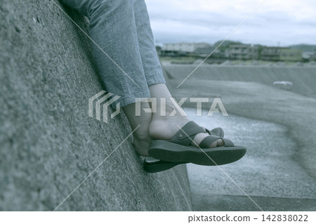 一張庫存照片，照片中一位女士的腳，赤腳和穿著涼鞋，坐在防波堤上。 142838022