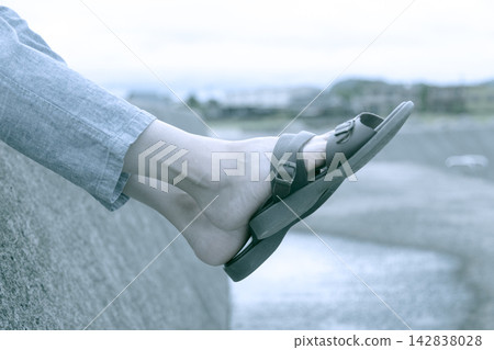 Stock photo of a woman's feet, barefoot and wearing sandals, sitting on a breakwater. Stock photo of a woman's feet, barefoot and wearing sandals, sitting on a breakwater. 142838028