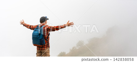 Man with backpack standing with raised arms in foggy mountains. Hiker in checkered shirt and cap enjoying freedom in misty landscape. Man with backpack standing with raised arms in foggy mountains. Hiker in checkered shirt and cap enjoying freedom in misty landscape. 142838058